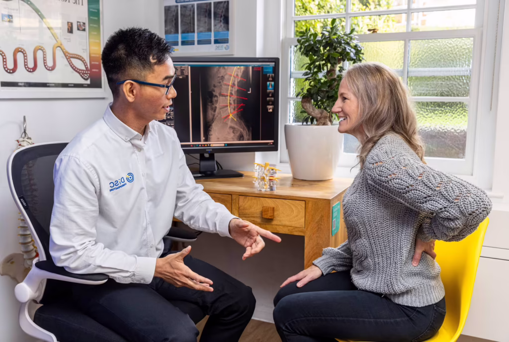 Chiropractor consulting with a woman experiencing lumbar lower back pain in a modern clinic, reviewing spinal X-rays during an initial chiropractic consultation.
