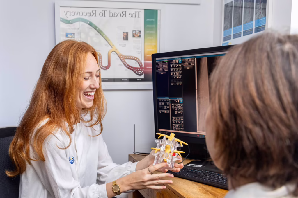 Chiropractor explaining a spinal disc model to a patient during a first consultation in Surbiton, with diagnostic imaging displayed on a computer screen in the background.